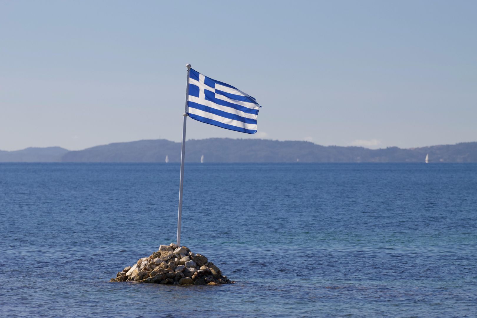 Die griechische Flagge weht über einer felsigen Insel vor der ruhigen Kulisse des Ionischen Meeres in Korfu, Griechenland.