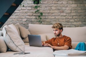 Ein Mann sitzt neben einer Couch und arbeitet am Laptop