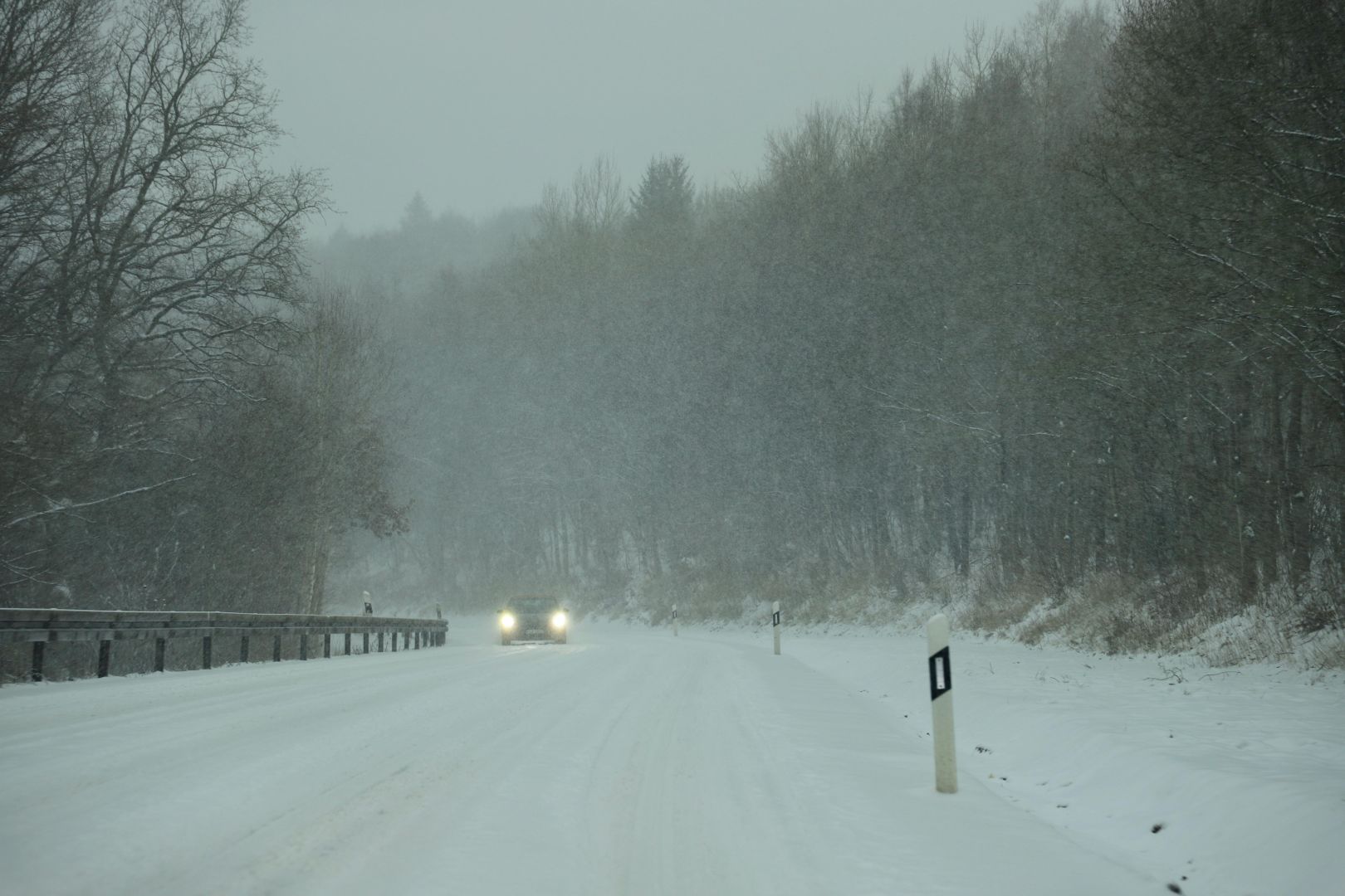 Ein Auto fährt während eines Schneesturms im Winter mit eingeschalteten Scheinwerfern auf einer schneebedeckten Straße.