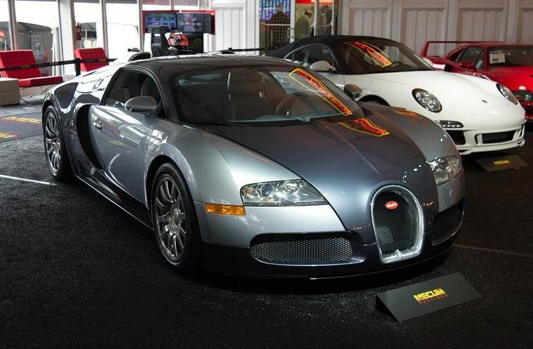 Silberner Bugatti Veyron bei einer Autoauktion in einer Ausstellungshalle, daneben ein weißer Porsche im Hintergrund.